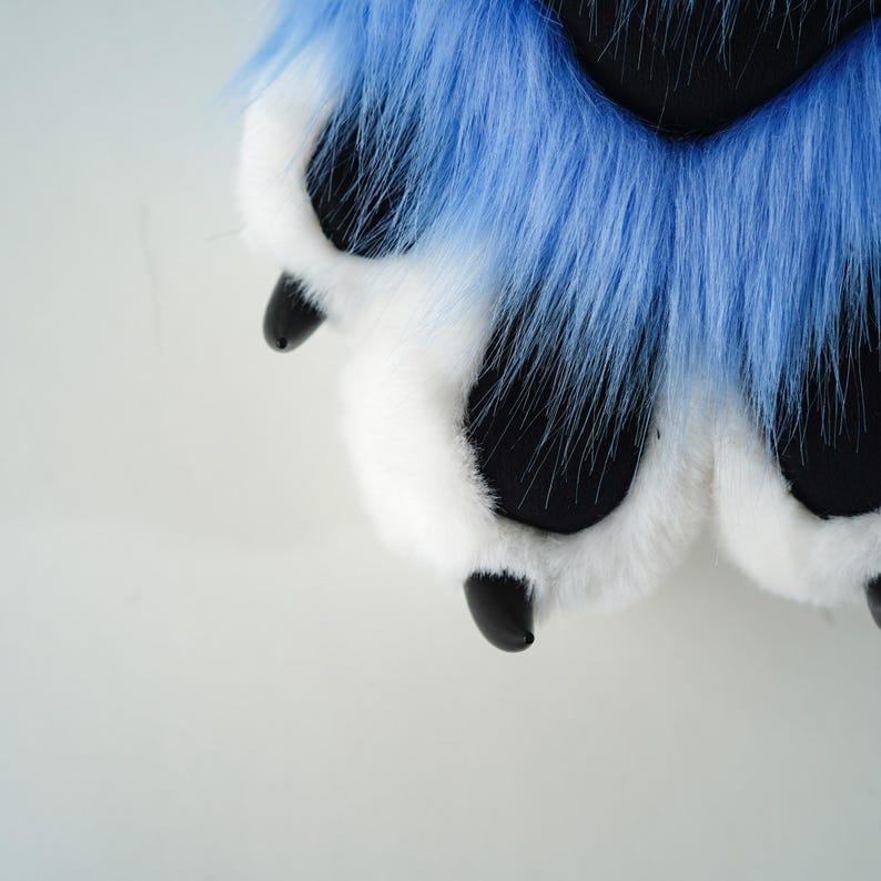 May include: Close-up of a furry paw with blue and white fur. The paw pads are black, and the claws are also black. The fur is long and fluffy, creating a soft and textured appearance. The overall design suggests a costume or plush toy.