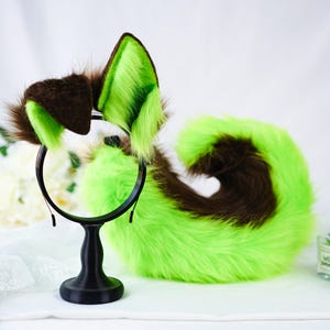Puede incluir: Un conjunto de accesorios de cosplay de piel, que incluye una diadema con orejas marrones y verde lima y una cola a juego. Las orejas y la cola tienen una textura esponjosa y est&aacute;n dise&ntilde;adas para parecerse a rasgos animales. Los accesorios se muestran en un soporte negro.