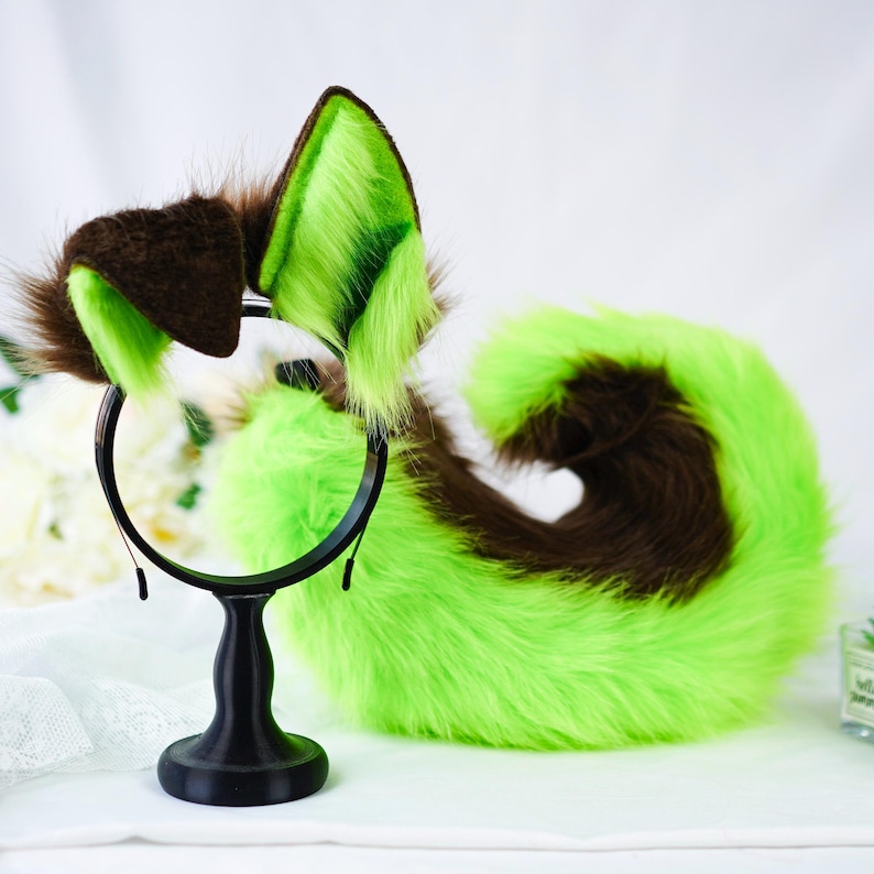 Puede incluir: Un conjunto de accesorios de cosplay de piel, que incluye una diadema con orejas marrones y verde lima y una cola a juego. Las orejas y la cola tienen una textura esponjosa y est&aacute;n dise&ntilde;adas para parecerse a rasgos animales. Los accesorios se muestran en un soporte negro.