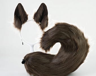 Puede incluir: Un conjunto de orejas de animal de piel sintética unidas a una diadema blanca y una cola larga y curva a juego. Las orejas y la cola presentan un pelaje marrón oscuro con reflejos blancos en los bordes, creando un aspecto texturizado.