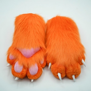 Può includere: Un paio di guanti a forma di zampa arancioni e pelosi con cuscinetti rosa e artigli bianchi. I guanti sono progettati per assomigliare a zampe di animali, con una consistenza morbida e soffice. Probabilmente fanno parte di un costume.