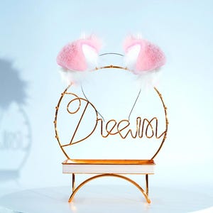May include: A pair of pink and white cat ears headband on a gold metal stand with the word "Dream" written in cursive.