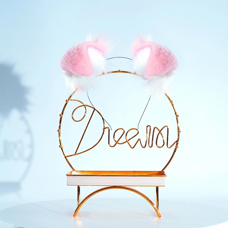 May include: A pair of pink and white cat ears headband on a gold metal stand with the word "Dream" written in cursive.