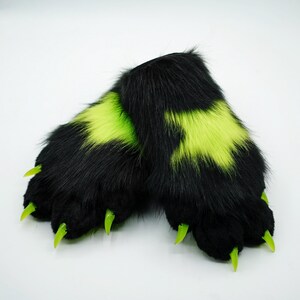 Puede incluir: Un par de patas peludas negras con garras verdes y detalles en verde ne&oacute;n. Las patas est&aacute;n dise&ntilde;adas para parecer patas de animales.