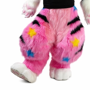 Könnte beinhalten: Flauschige rosa Hose mit schwarzen tigerartigen Streifen und verstreuten gelben und blauen Sternenmustern. Das Kleidungsstück umfasst weiße Plüschpfoten mit rosa Ballen an den Füßen und wird mit einem schwarzen Oberteil kombiniert.