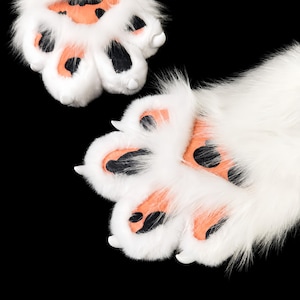 May include: A pair of plush white faux fur animal paws featuring soft peach-colored paw pads with black spots and small white claws. The textured fur is long and thick, covering the entire surface of the paws, which are displayed against a black background.
