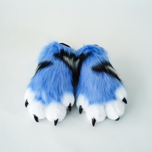 May include: A pair of furry paw-shaped slippers. The slippers are primarily light blue with black stripes and white paw pads. The claws are black. The slippers are designed to resemble animal paws.