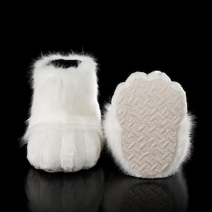 Op de afbeelding: Een paar witte imitatiebont pantoffels in de vorm van dierenpoten. Eén pantoffel is van voren te zien, met de pluizige textuur en afgeronde neus, terwijl de andere de gestructureerde, gebroken witte rubberen zool aan de onderkant laat zien.