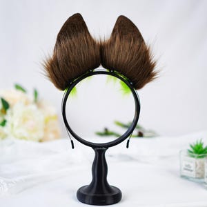 Puede incluir: Una diadema con orejas de gato de piel sint&eacute;tica marr&oacute;n con una montura negra. Las orejas son esponjosas y erguidas, con un acento verde claro. La diadema se muestra sobre un soporte negro sobre un fondo blanco.