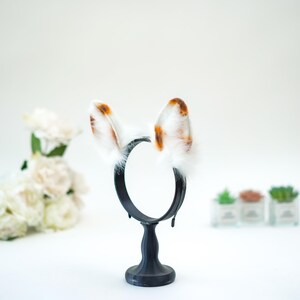 May include: A black headband with white and brown spotted bunny ears. The ears are fluffy and stand upright. The headband is displayed on a black stand against a white background. Decorative flowers and small potted plants are in the background.