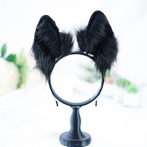 May include: Black furry cat ears headband on a black stand. The headband is made of a black elastic band with two black furry cat ears attached.