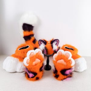 May include: A set of orange and black tiger-themed fursuit parts, including a tail, paws, and a headpiece with ears. The headpiece has a white and black face with pink accents.