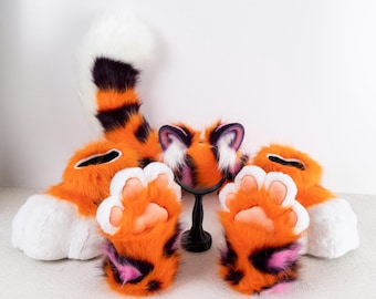 May include: A set of orange and black tiger-themed fursuit parts, including a tail, paws, and a headpiece with ears. The headpiece has a white and black face with pink accents.