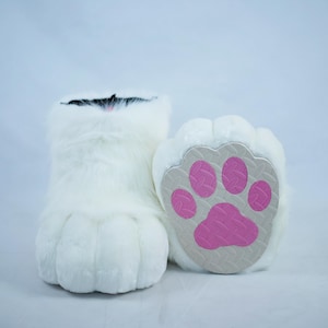 Op de afbeelding: Witte, harige kattenpoot-pantoffels met een roze pootprint op de onderkant.
