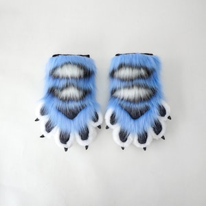 May include: A pair of furry paw gloves in light blue, white, and black. The gloves have a paw-like design with black claws and are likely part of a costume or cosplay accessory.