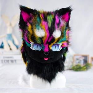 May include: A colorful cat fursuit head with black fur, pink, green, yellow, and white markings. The fursuit has large blue eyes and a pink tongue sticking out.