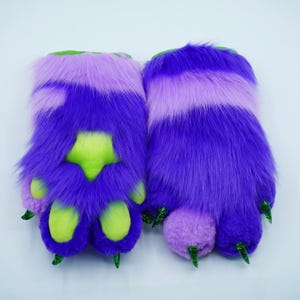 May include: A pair of purple and green furry paw hand puppets with green claws. The paws have a light purple stripe and a green paw pad.