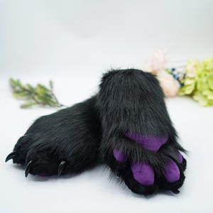 Könnte beinhalten: Ein Paar schwarze, pelzige Handpuppen mit violetten Pfoten. Die Puppen liegen auf einer weißen Oberfläche.