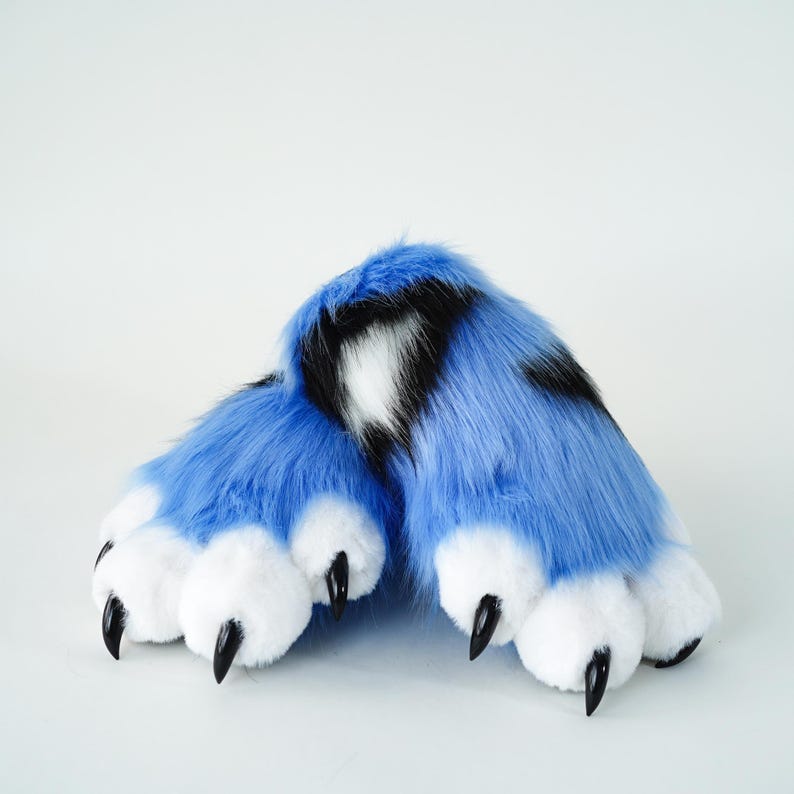 May include: A pair of blue faux fur paws with black and white markings. The paws have white pads and black claws. The furry costume feet are designed to resemble animal paws.
