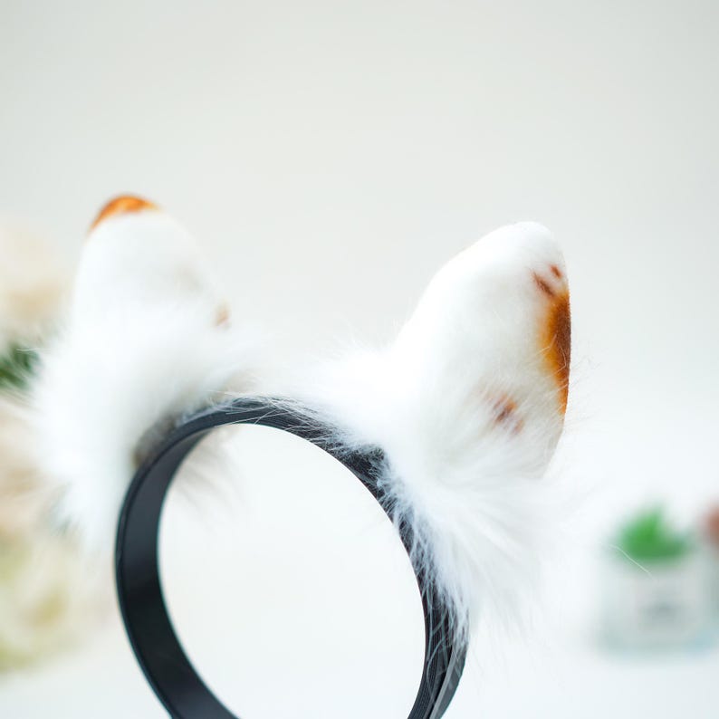 May include: A black headband with attached white cat ears. The ears are fluffy with brown spots and a soft, furry texture. The headband is made of a smooth, solid material.