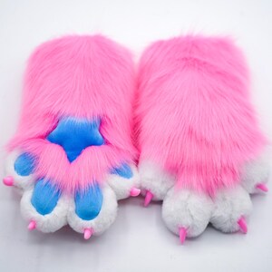 Puede incluir: Un par de calentadores de manos de pata peluda rosa con almohadillas de pata azules y garras rosas.
