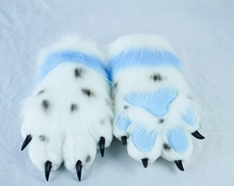 Puede incluir: Un par de guantes de pata blancos y peludos con manchas negras y garras negras. Los guantes tienen un acento azul claro alrededor de la muñeca y almohadillas de pata azul claro.