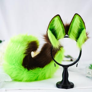 Puede incluir: Un conjunto de accesorios de cosplay con una diadema con orejas de zorro verdes y marrones y una cola a juego. Las orejas son de color verde brillante con puntas marrones, y la cola es verde y marr&oacute;n. Se muestra sobre un soporte negro.