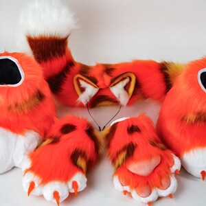 以下が含まれることがあります： レッサーパンダをテーマにした、毛むくじゃらのコスチュームセット。セットには、尻尾、耳付きヘッドバンド、足の形をした手袋が含まれています。尻尾は茶色と白のストライプで、手袋にはオレンジ色の爪が付いています。