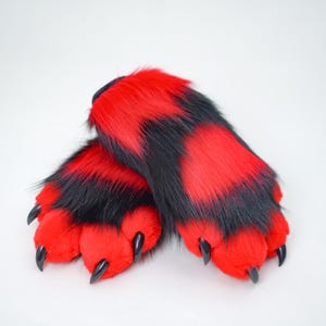 May include: A pair of furry paw-shaped costume feet in red and black. The paws have black claws and are designed to resemble animal feet. The fur is a mix of red and black, creating a striped pattern.