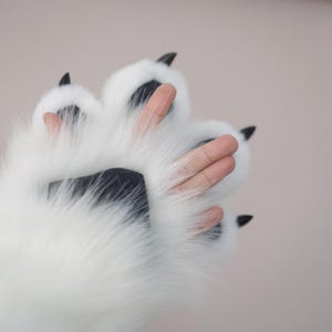 May include: A close-up of a white faux fur paw with black pads and claws. The paw has visible fingers and is set against a soft, neutral background. The fur is fluffy and the claws are sharp.