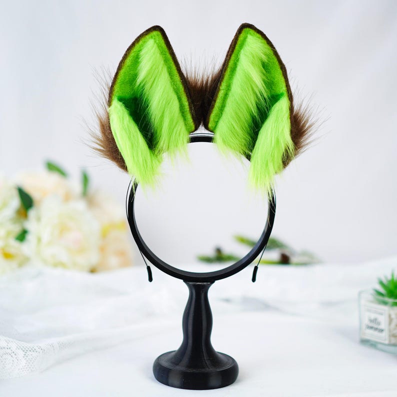 Puede incluir: Una diadema con orejas de animal de piel sint&eacute;tica. Las orejas son marrones con pelo interior verde brillante. La diadema es negra y las orejas est&aacute;n montadas sobre un soporte negro.