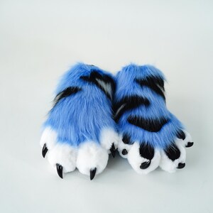 May include: A pair of blue and black furry paw-shaped slippers. The slippers have white paw pads and black claws. The fur is a vibrant blue with black stripes. The slippers are designed to resemble animal paws.