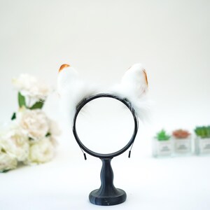 May include: A black headband with fluffy white cat ears, tipped with brown. The ears are attached to a black circular frame, presented on a black stand. The background is white with blurred floral and plant accents.