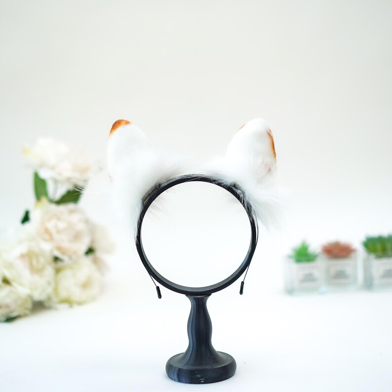 May include: A black headband with fluffy white cat ears, tipped with brown. The ears are attached to a black circular frame, presented on a black stand. The background is white with blurred floral and plant accents.