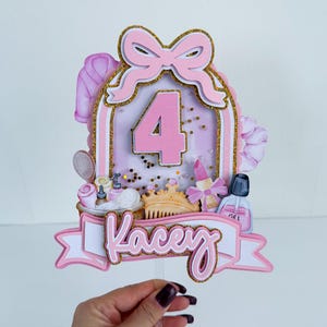 May include: A pink and gold glitter cake topper with the number 4, a bow, and the name "Kacey". The topper features beauty-themed elements like a comb, nail polish, and a miniature sink. The overall design is festive and celebratory.