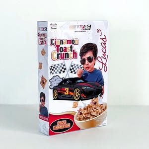 May include: Cinnamon Toast Crunch cereal box featuring a cartoon illustration of a race car and a child wearing sunglasses. The box has the text "Cinnamon Toast Crunch" and "2023 Champion". The cereal is in a bowl.
