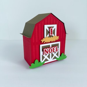 May include: A red paper barn-shaped box with a brown roof. The box has a white door with the word "NOEL" and the number "1" on it. Green grass-shaped cutouts are at the bottom of the box.