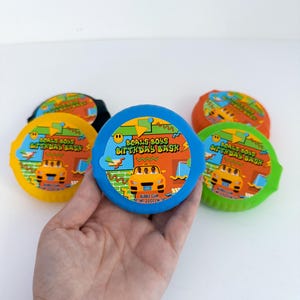 May include: A hand holding a blue, round container of bubble gum with the text "BOALS BOUS BIRTHDAY BASK" and a cartoon car graphic. Other containers in yellow, orange, and green are visible.