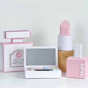 May include: Pink and white cardboard boxes shaped like perfume bottles and lipstick, with the name "Shyla Soul" on one of the boxes. The boxes are arranged on a white surface.