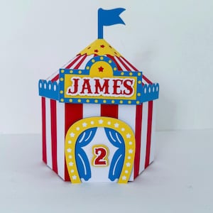 May include: A colorful circus-themed box with red and white striped sides. The top is yellow with red stars and a blue flag. The box has the name "JAMES" and the number "2" on it.