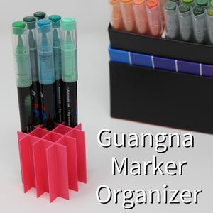 Puede incluir: Un organizador de rotuladores rosa contiene varios rotuladores con tapas verdes y azules. Una caja negra con una muestra de color y rotuladores está en el fondo. Se muestra el texto "Guangna Marker Organizer".