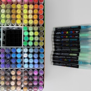 A collection of colorful art markers, neatly organized in a white storage container. The markers are arranged by color, with a wide range of hues visible. A stack of additional markers is to the right. The image showcases a variety of art supplies.