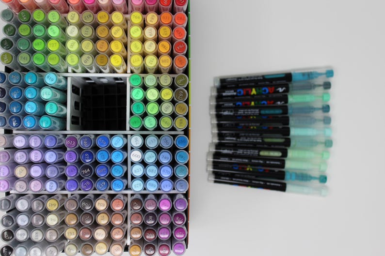 A collection of colorful art markers, neatly organized in a white storage container. The markers are arranged by color, with a wide range of hues visible. A stack of additional markers is to the right. The image showcases a variety of art supplies.