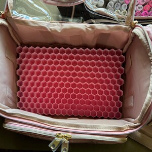 May include: Open pink makeup bag with gold zippers and a clear top. Inside, a pink honeycomb-shaped organizer is visible. The bag is designed to hold makeup and beauty supplies, offering organized storage.