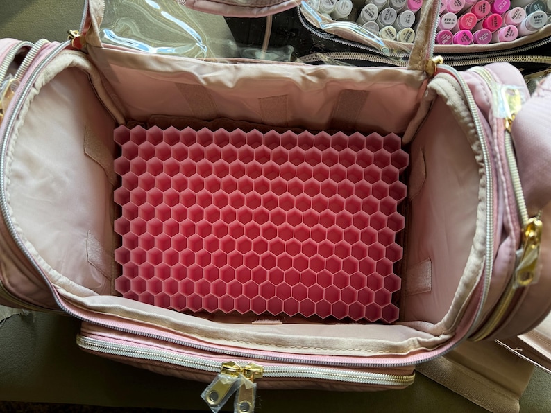 May include: Open pink makeup bag with gold zippers and a clear top. Inside, a pink honeycomb-shaped organizer is visible. The bag is designed to hold makeup and beauty supplies, offering organized storage.