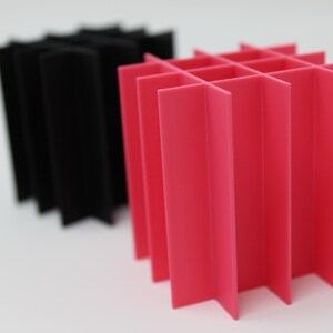 Two geometric, abstract sculptures. One is black, the other is bright pink. Both are constructed from folded, intersecting planes, creating a grid-like structure. The sculptures are set against a plain white background.