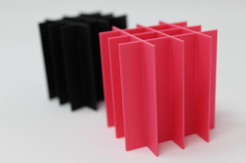 Two geometric, abstract sculptures. One is black, the other is bright pink. Both are constructed from folded, intersecting planes, creating a grid-like structure. The sculptures are set against a plain white background.
