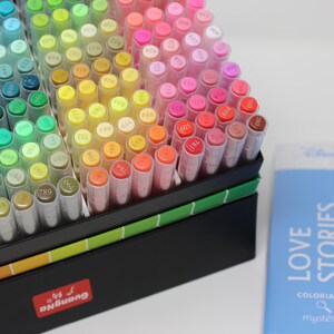 A collection of colorful art markers in a black box with a rainbow gradient. The markers are arranged by color, ranging from blues and greens to yellows, oranges, and pinks. A blue book with the title "LOVE STORIES" is also visible.