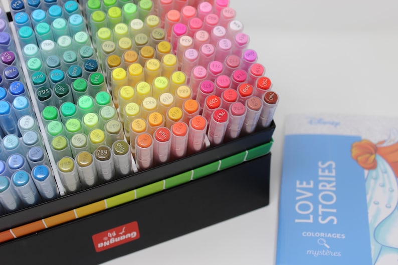 A collection of colorful art markers in a black box with a rainbow gradient. The markers are arranged by color, ranging from blues and greens to yellows, oranges, and pinks. A blue book with the title "LOVE STORIES" is also visible.