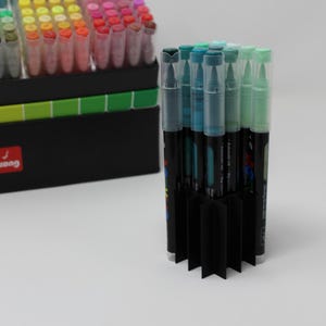 A collection of colorful art markers with clear caps, arranged in a black organizer. The markers are in various shades, including blue, green, pink, and yellow. A second set of markers is visible in the background.